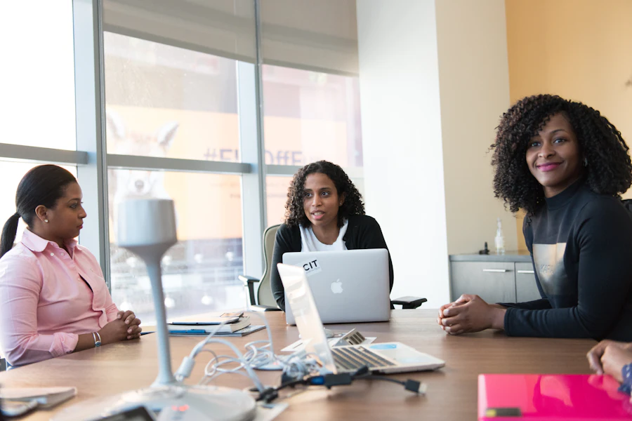 Three professional women in conversation around a table with a laptop in a bright office