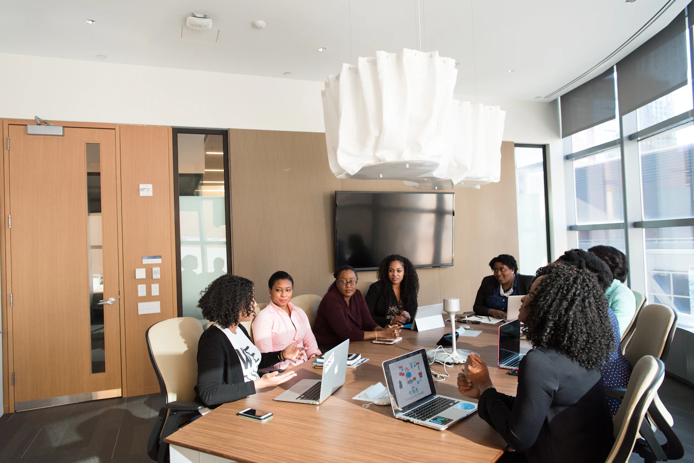 A professional team, largely Black women, meeting around a wooden table with laptops