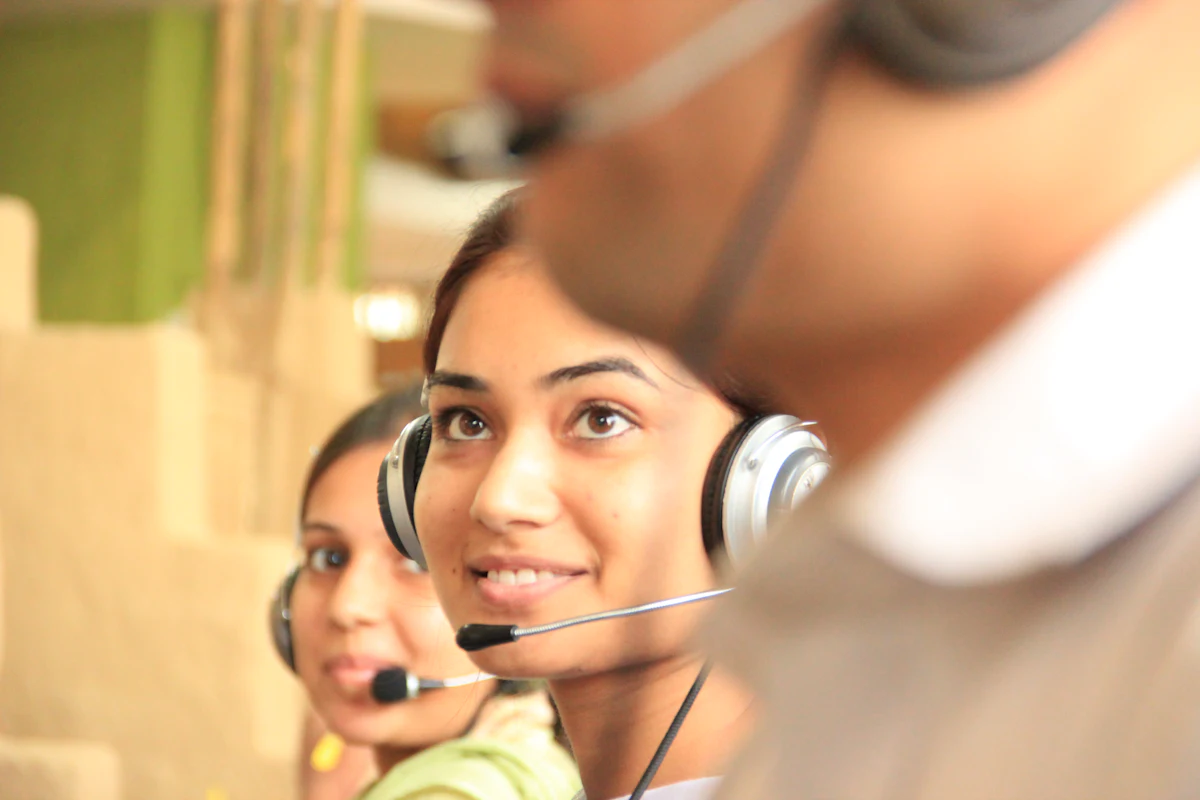 Smiling customer support agent wearing a headset with a colleague in the foreground of a contact centre