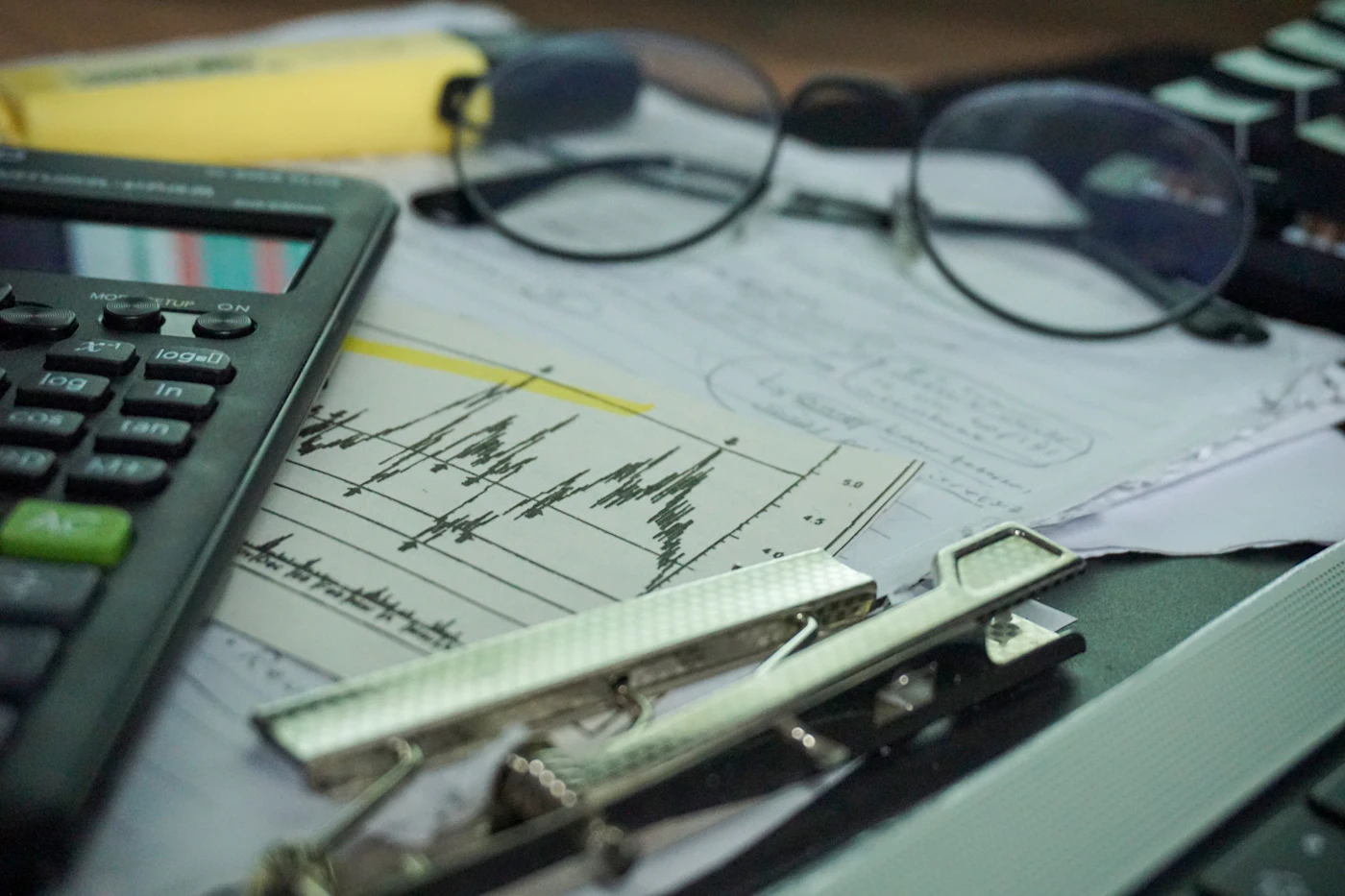 A desk with a calculator, eyeglasses, clipboard and printed financial charts, representing bookkeeping work